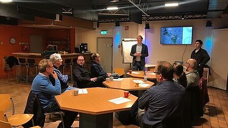 Twee mannen geven een presentatie aan 7 mensen aan tafels