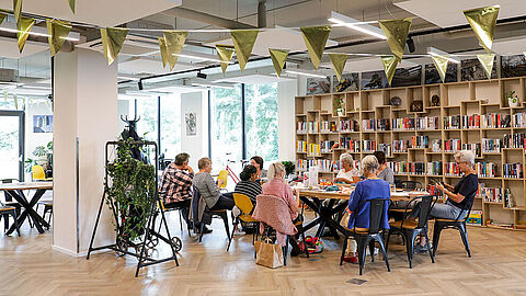 Ouderen zitten aan een ronde tafel met gouden vlaggetjes erboven en een boekenkast op de achtergrond