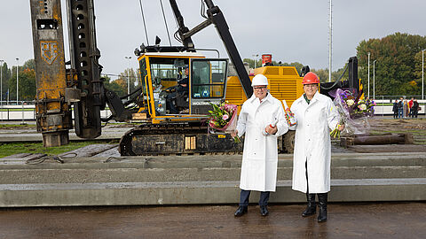 Wethouder John van Engelen en levenspartner Brigit Rams van Michel van den Hoven staan met bloemen voor de bouwplaats