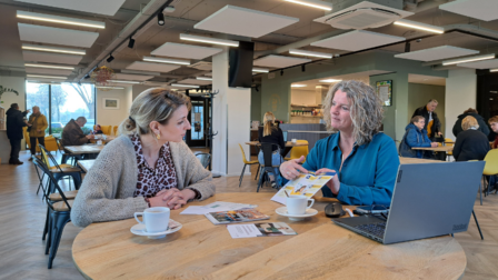 Twee vrouwen aan een tafel, de ene vrouw wijst iets aan op een folder